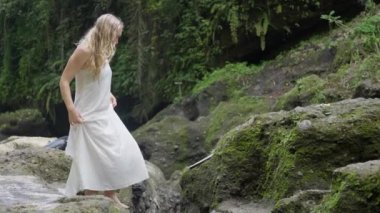 Güzel İskandinav bir kadın, dalgalı uzun sarı saçlı, İskandinav bir kadın, güzel beyaz elbiseli, yeşil yosunlarla kaplı kayalar üzerinde yürüyor. Ağır çekim. Yan görünüm Yüksek kalite 4k