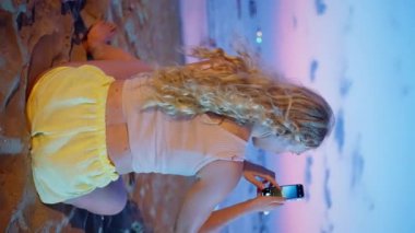 Genç sarışın kadın gün batımında sakin bir şekilde tropikal sahilde oturuyor. Yalnız kız yazın güzel kumsala bakıyor. Arka planda güneş ve okyanus var. Dikey görünüm