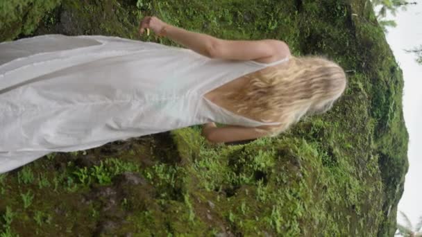 一个金发碧眼的漂亮女人 一个金发碧眼 身穿白色连衣裙的女孩 站在岩石中央 在阳光灿烂的一天观看瀑布 慢动作纵向观点 — 图库视频影像