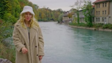 Sarışın, uzun saçlı ve yeşil gözlü güzel bir kadının yakın çekimi. Köydeki bir dağ nehrinin arka planında ceketi ve beyaz şapkasıyla İskandinav görünümlü çekici bir kadın. Yavaş çekim