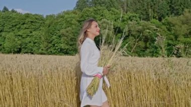 Beyaz elbiseli güzel Ukraynalı kadın elinde buğday başaklarıyla bir buğday tarlasında yürür arka planda mavi bir gökyüzü. Yavaş çekim.