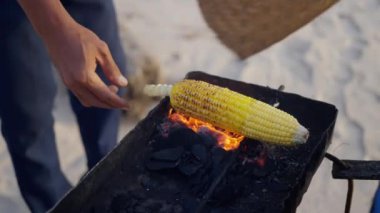 Bir insan, el ve aracı doğaçlama bir ızgara olarak kullanarak, sakin bir plaj etkinliği için, bir palmiye ağacının yanındaki koçanda mısır pişiriyor. Ağır çekim. Tayland. Pattaya..