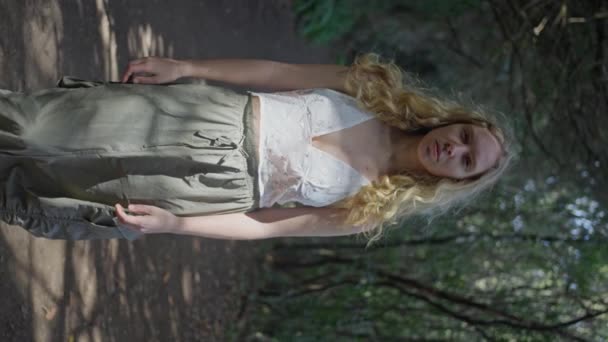 白いトップと緑のズボンを着た長い髪の女性が森の中の木の間に立っており 自然の中で人間の体の美しさを示しています スローモーション 垂直ビュー 4Kについて — ストック動画