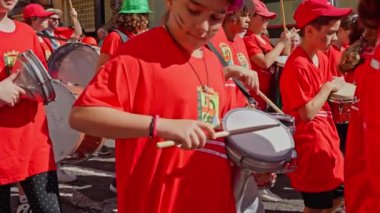 Bir geçit töreninde davul, zar ve rüzgar enstrümanları çalan kırmızı tişörtlü bir grup çocuk müzisyen. Müzikli eğlence etkinliği. Tenerife 18 03 2024. Ağır çekim. Yüksek kalite 4k görüntü