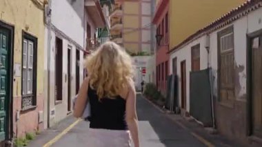 Beyaz etekli kız dar bir yolda yürüyor, asfalt güneş ışığı altında parlarken bitkilerin ve binaların yanından geçiyor. Arkadan bak. Zaman turları. Tenerife 14 02 2024