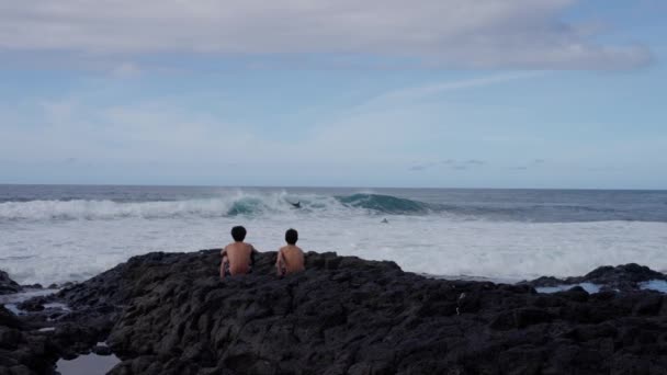 两个男孩坐在海边的岩石上 凝视着海浪冲击着海岸 天空布满了云彩 地平线伸展在水面之上 慢动作 Tenerife — 图库视频影像