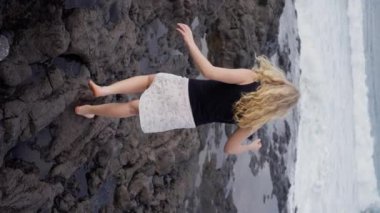 Siyah ve beyaz etekli yetişkin kız Tenerife adasında kayalık bir plaj boyunca yürüyor. Mutlu görünüyor, rahatlamanın tadını çıkarıyor, huzurlu bir ortamda eğleniyor. Ağır çekim. Dikey görünüm. 4k