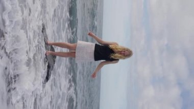 Yetişkin bir kız bir kayanın üzerinde duruyor, sıvı bir okyanusun yanında kumlu bir plaj, üzerinde gökyüzü ve sonsuz suya bakıyor, resim gibi bir manzara yaratıyor. Ağır çekim. Dikey video. Yüksek kalite 4k