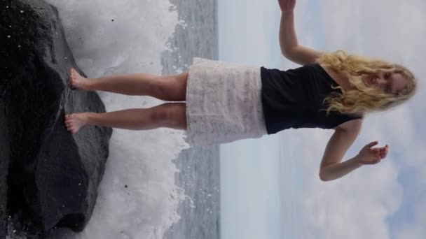 一个女人站在岩石上 一个沙滩上 旁边是一个流动的海洋 天空在她的上方 她看着无尽的水 创造了一个风景如画的场景 慢动作垂直录像 高质量4K — 图库视频影像