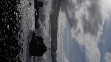 Güneş, buz gibi gri bulutların arasından sakin bir manzaraya bakıyor. Dalgalar siyah kum kıyısına çarpar. Siyah ve beyazın zıtlığı doğanın güzelliğini vurgular. Yavaş Hareket Dikey Görünümü