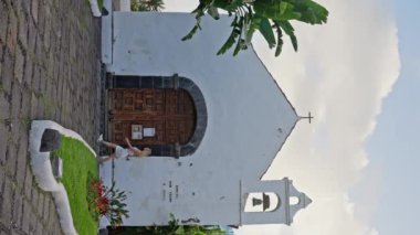 Binanın tepesinde çan kulesi olan küçük beyaz bir kilise ağaçlar ve yakınlardaki evlerle çevrili açık mavi gökyüzünün altında yemyeşil çimlerin arasında duruyor. İspanya, Tenerife. Dikey oryantasyon. 4k
