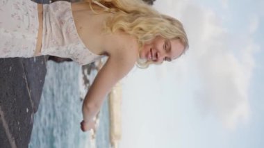 Açık mavi gökyüzünün altında okyanus kenarında beyaz elbiseli bir kız. Arkasını dönüp gülüyor, seviniyor ve okyanusu işaret ediyor. Tenerife İspanya. Ağır çekim. Dikey görünüm. Yüksek kalite 4k görüntü