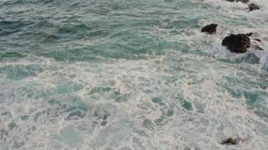 Dev okyanus dalgalarının yukarıdan aşağıya yukarıdan görünüşü büyük kaya taşlarıyla dolu Tenerife adasının boş kıyılarına çarpıyor ve köpürüyor. Altın plajın derin mavi okyanus suyuyla buluşmasının kuş bakışı görüntüsü.