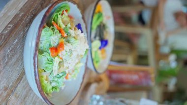 İki kase salata, sofra takımı ve cam tabaklarla çevrili, ahşap bir masanın üzerine konur. Renkli malzemeler güzel bir sanat gösterisi yaratıyor.