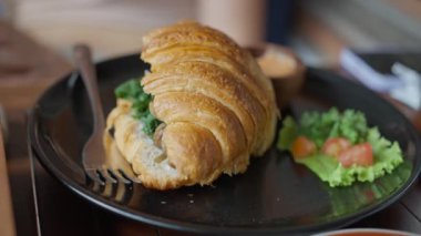 Pürüzsüz siyah bir tabakta servis edilen kruvasan sandviçi ve şık bir sunum için çatal. Sandviç taze ürünler ve fırınlanmış gıdalarla yapılır. Lezzetli bir yemek için mükemmeldir.