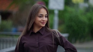 Kendine güvenen, gülümseyen, koyu renk tişörtlü, çevresi yeşilliklerle çevrili genç bir kadın. Yavaş çekim