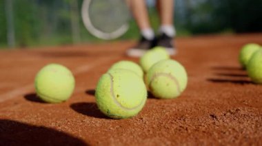 Şort giyen bir adam tenis toplarıyla çevrili bir tenis sahasında duruyor ve maça hazırlanıyor. Çitlerle çevrili kil kort. Yavaş çekim 4k