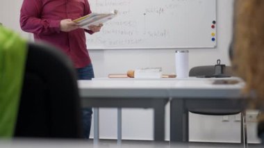 Öğretmen ayakta ve ön planda beyaz tahtası ve masası olan bir sınıfta kitap okuyor..