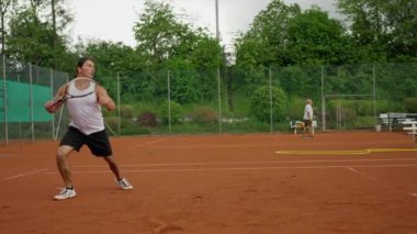 Bir adam kil sahada tenis oynayarak, atletizm ve tutku sergileyerek yetenek ve adanmışlık gösteriyor. Yavaş çekim
