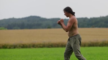 Üstsüz bir adam, sakin bir çimenlikte kırsal bir manzaraya karşı dövüş sanatları yapıyor. Yavaş çekim 4k