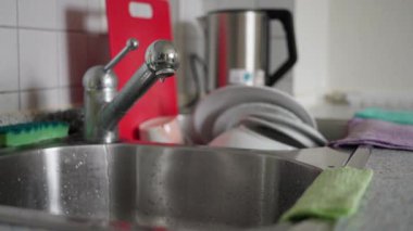 Mutfak lavabosunun yakından çekilmiş bir fotoğrafı. Yanında damlayan bir musluk ve dikkatlice istiflenmiş bulaşıklar var. Yavaş çekim