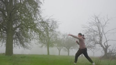 Sonbaharın sonlarında sabah sisli tarlasında kickboks antrenmanı yapan bir adam. Ağır çekim. Sisli bir alanda enerjik bir duruş içinde olan bir kişi, doğada yaratıcılık ve dinamik ifade sergiliyor.