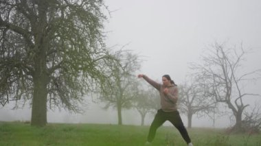 Sonbaharın sonlarında sabah sisli tarlasında tekvando antrenmanı yapan bir adam. Ağır çekim. Sisli bir alanda enerjik bir duruş içinde olan bir kişi, doğada yaratıcılık ve dinamik ifade sergiliyor.