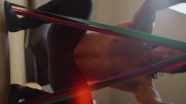 Formda bir adam, evindeki spor salonunun rahatlığında direnç bantlarını kullanarak çeşitli bacak egzersizleri yapıyor. 4K görüntü. Dikey video