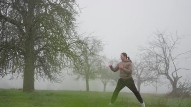 Sonbaharın sonlarında sabah sisli tarlasında kickboks antrenmanı yapan bir adam. Yavaş çekim 4K. Sisli bir alanda enerjik bir duruş içinde olan bir kişi, doğada yaratıcılık ve dinamik ifade sergiliyor.