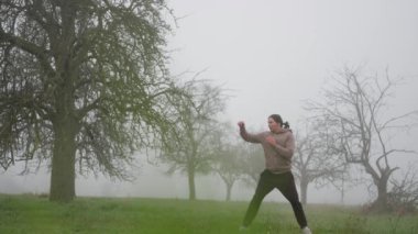 Sonbaharın sonlarında sabah sisli tarlasında kickboks antrenmanı yapan bir adam. Ağır çekim. Sisli bir alanda enerjik bir duruş içinde olan bir kişi, doğada yaratıcılık ve dinamik ifade sergiliyor.