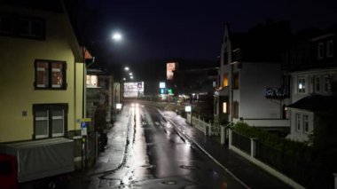 Geceleri sessiz bir cadde, ıslak kaldırımlar sergileniyor ve sakin bir şehir ortamında aydınlatılmış binalar..