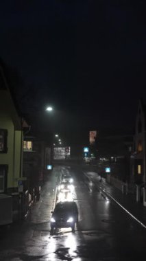 Yağmurlu bir gecede, sakin bir cadde manzarası parlayan ışıklar ve hareket eden bir araba havayı yakalar.