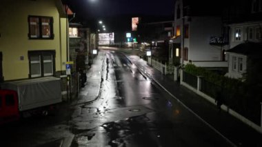 Huzurlu bir gecede, sokak lambaları ve binaların ışığında parıldayan ıslak bir caddenin güzel bir manzarasını görün.