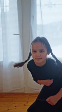 Büyüleyici siyah elbiseli genç bir kız örgülü saçlarıyla neşeli bir şekilde poz veriyor, neşeli bir ruh yayıyor. Çocuk dans eder ve gülümser.