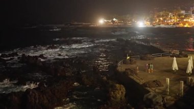 Okyanusun sakin bir gece manzarası. Kayalara çarpan dalgalar ve arka planda şehir ışıkları..