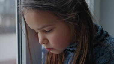 Küçük bir çocuk düşünceli bir şekilde pencereden dışarı bakıyor. Kapalı bir ortamda çeşitli düşünceleri derin bir şekilde yansıtıyor.