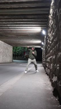Gece köprünün altında kapüşonlu tren giyen bir adam, sokakta atlet kutuları. Enerjik bir figür, geceleri şehir mimarisi altında sokak tarzı gösteriler yaparak dinamik hareketler sergiliyor. yavaş çekim