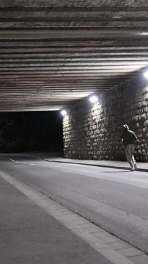 Gece köprünün altında kapüşonlu tren giyen bir adam, sokakta atlet kutuları. Enerjik bir figür, geceleri şehir mimarisi altında sokak tarzı gösteriler yaparak dinamik hareketler sergiliyor. Ağır çekim. 4k