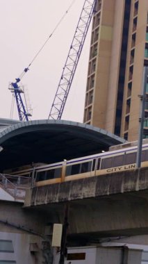 Yükseltilmiş bir tren hızla modern bir istasyona yaklaşıyor ve arka planda açık bir şekilde inşaat devam ediyor.