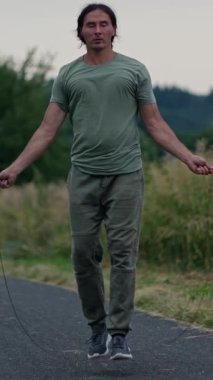 Atletik bir adam, alacakaranlıkta doğa tarafından çevrelenmiş güzel, manzaralı bir yolda enerjik bir şekilde ip atlıyor.