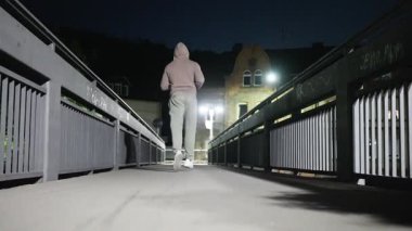 Uzun bir köprüde, loş sokak lambalarıyla aydınlatılan yalnız bir koşucu, geceleri huzurlu bir şehir ortamı yaratır.