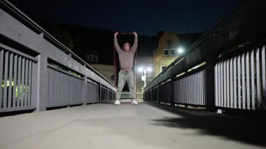 Bir insan, gece vakti köprüde ayakta dururken direnç bantları kullanarak güçlü bir çalışma yapıyor.