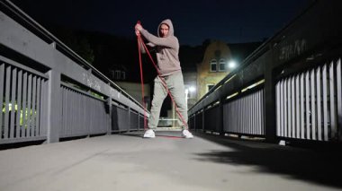 Bir adam, dayanıklılığını ve uygunluğunu vurgulamak için geceleri köprüde direnç bandı egzersizleri yapıyor. Yüksek kalite 4k görüntü