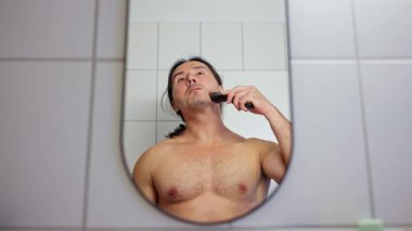 Bir adam aynanın önünde sakalını tımar ediyor, günlük bakımını yapıyor ve kişisel sağlığını destekliyor.