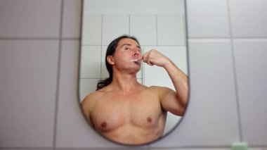 Bir adam dişlerini banyoda fırçalar, aynada canlı bir şekilde yakalanır, günlük hijyenini gösterir.
