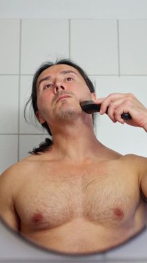 Bir adam aynanın önünde sakalını tımar ediyor, günlük bakımını yapıyor ve kişisel sağlığını destekliyor.