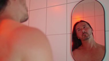 Bir adam aynanın önünde saçını şekillendiriyor. Arka planda canlı kırmızı bir ortam var.