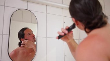 Bir adam banyo aynasında tıraş olur, bakımını ve bakımını iyi korunmuş bir görünüş için ön plana çıkarır.