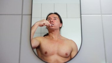 Bir adam dişlerini banyoda fırçalar, aynada canlı bir şekilde yakalanır, günlük hijyenini gösterir. Ağır çekim. Yüksek kalite 4k görüntü