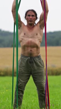 Üstsüz bir adam dışarıda direnç bandı egzersizi yapar. Güç ve esneklik gösterir..